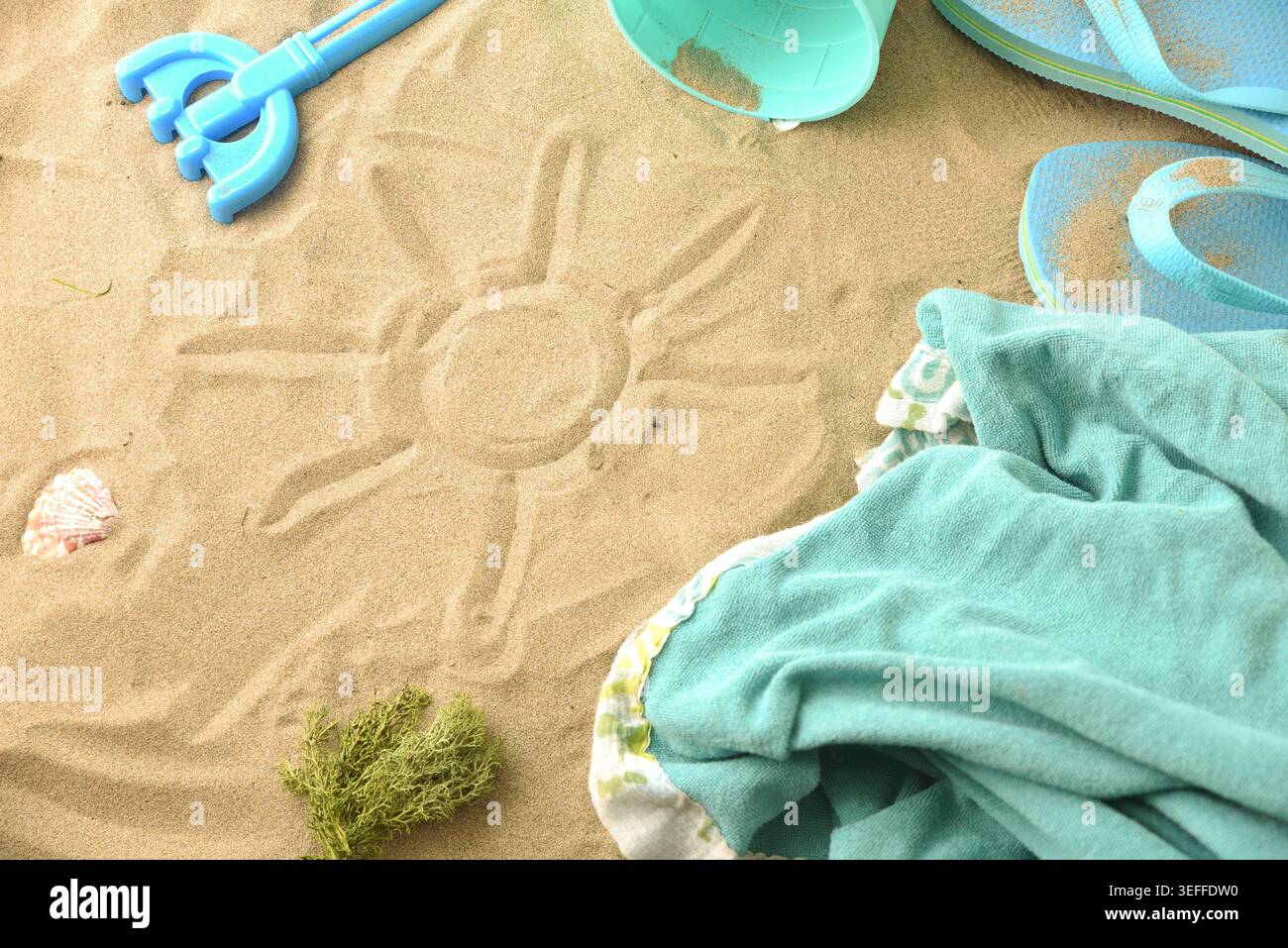 Simbolo del sole disegnato a mano sulla spiaggia sabbiosa circondato da accessori estivi, infradito, asciugamano, conchiglie e secchio in piena luce del giorno. Foto Stock