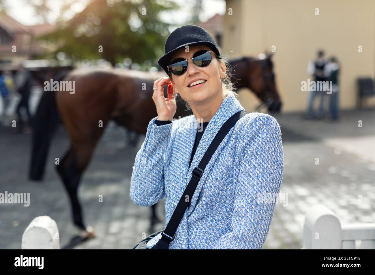 Elegante donna sorridente che indossa occhiali da sole e berretto vicino ai cavalli nella stalla di campagna. Donna elegante che gode di una giornata di sole all'aperto con relax Foto Stock