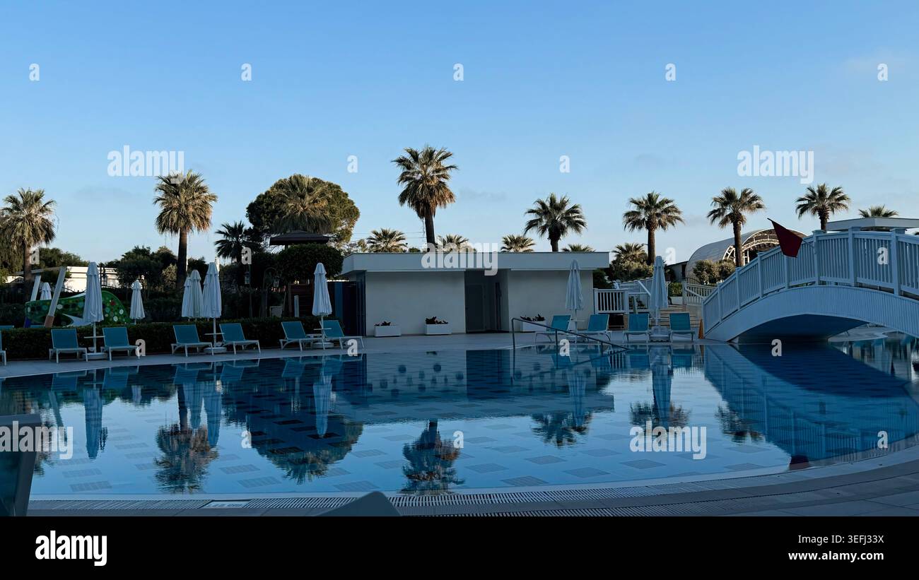 Area piscina esterna con palme e sedie a sdraio, che riflettono il cielo blu e le nuvole, caratterizzata da un ponte bianco e da una moderna struttura a bordo piscina Foto Stock