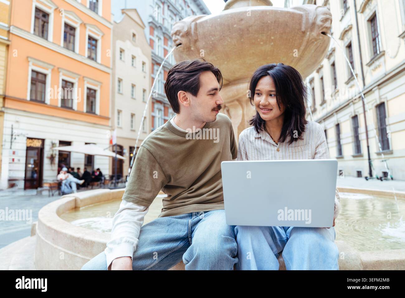 Felice giovane coppia che usa un computer portatile insieme mentre si siede accanto alla fontana nella città europea Foto Stock