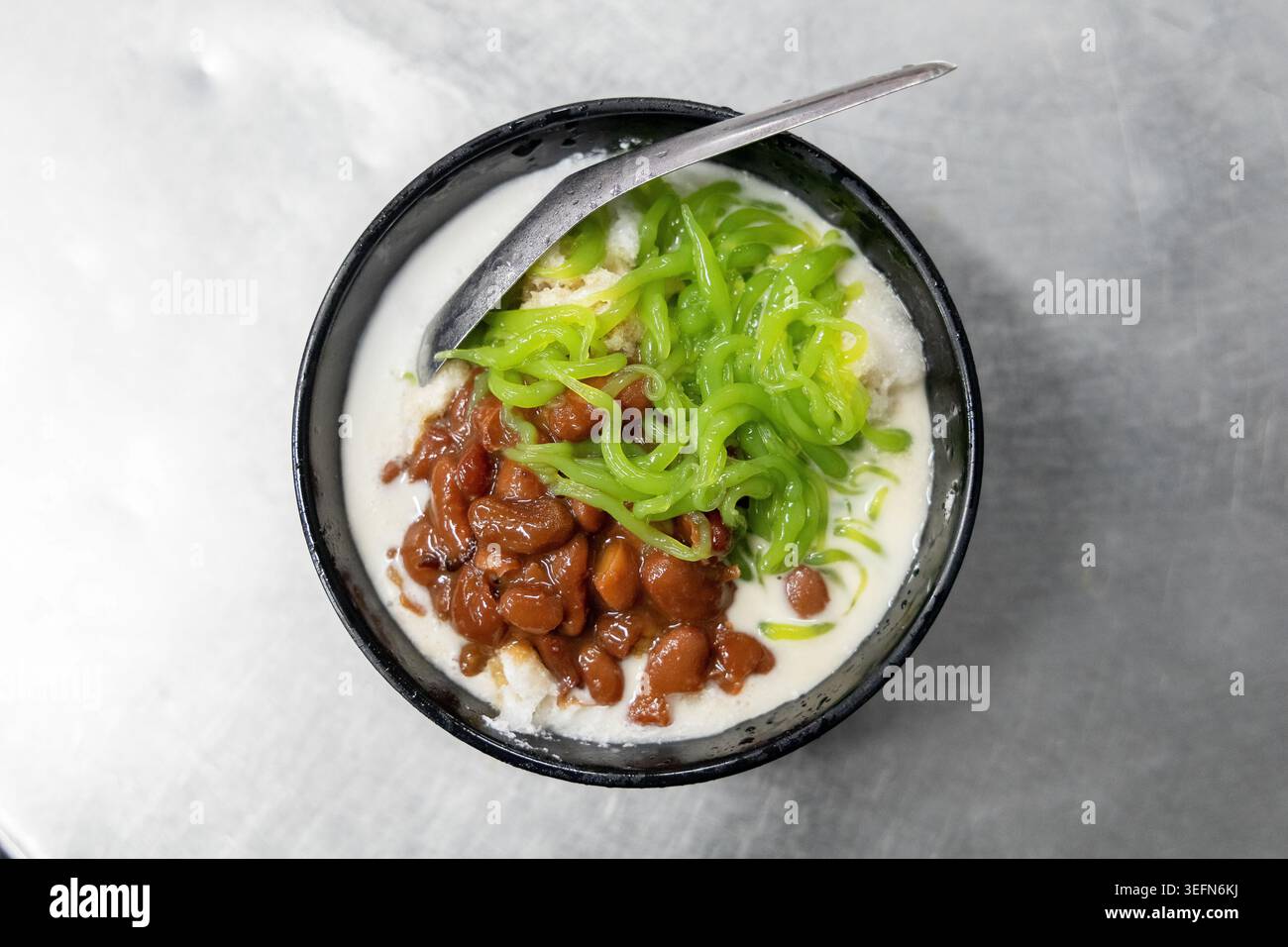 Una vista dall'alto di una ciotola di dolce cendol malese, con filamenti di gelatina di pandan verde brillante, fagioli rossi e ricco latte di cocco, servito in una ciotola di ceramica scura su una superficie di marmo grigio. Il classico dessert cattura i colori vivaci della cultura gastronomica dei falchi di Penang. Foto Stock