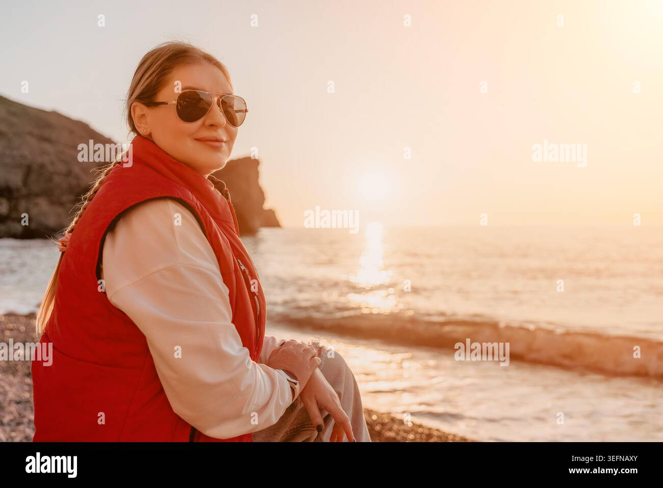 Donna al tramonto sulla spiaggia rocciosa in gilet rosso e occhiali da sole durante la tranquilla ora d'oro con spazio fotocopie Foto Stock