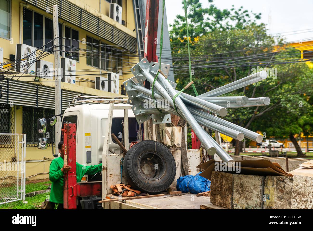 Questa immagine mostra una scena di costruzione con un camion che trasporta tubi metallici sollevati da una grande gru, sfondo urbano, operaio edile in sicurezza Foto Stock