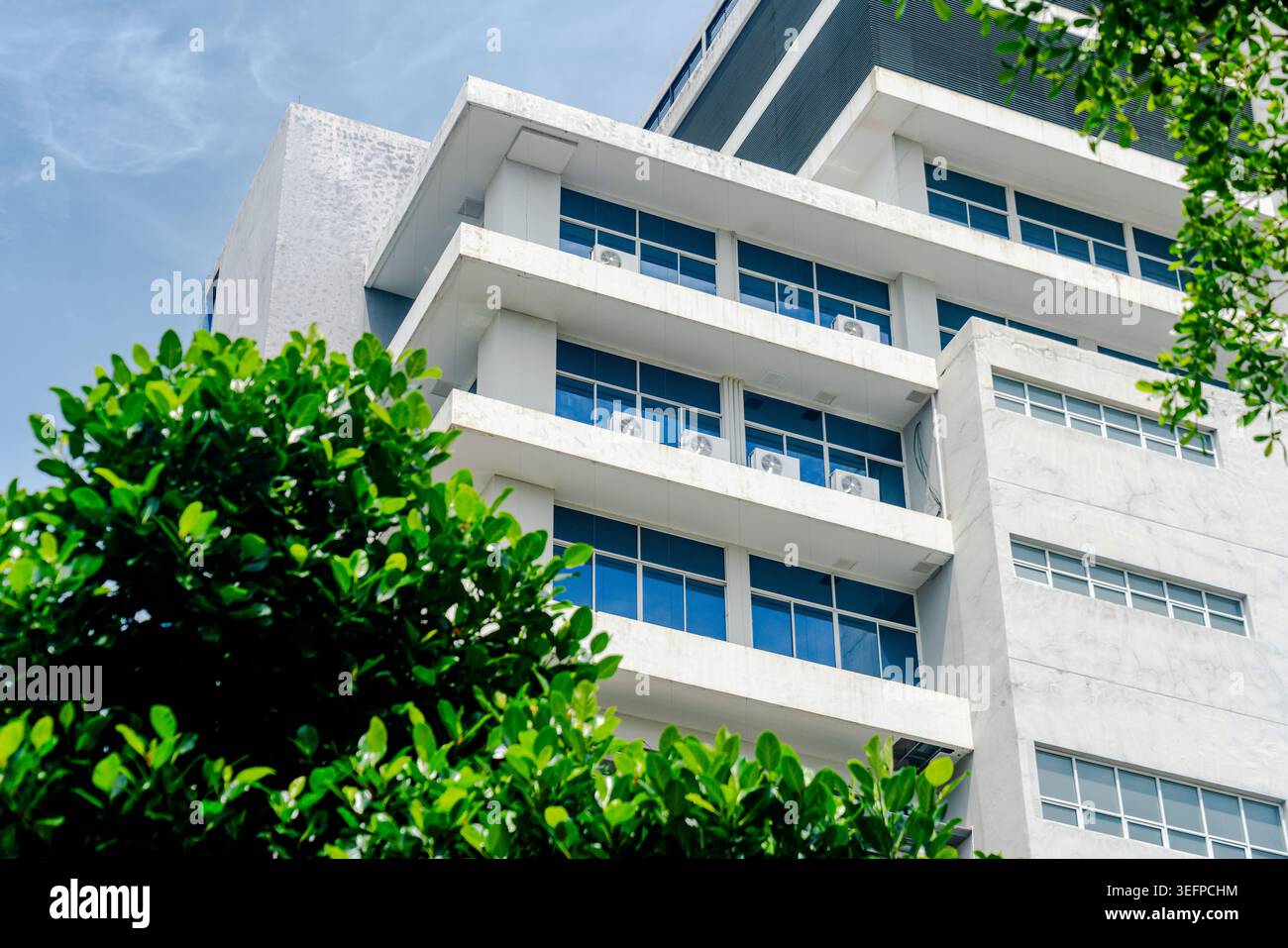Un moderno edificio residenziale bianco a più piani caratterizzato da grandi finestre in vetro, balconi e un design elegante in mezzo a lussureggiante vegetazione verde, sotto un luminoso edificio Foto Stock