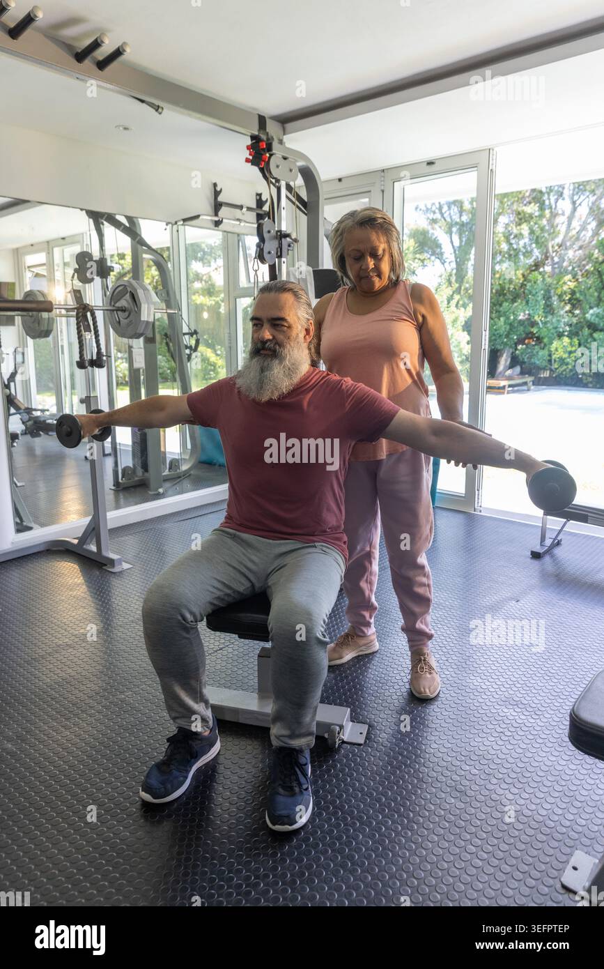 Coppia senior che si esercita a casa, uomo che solleva manubri sulla panchina, donna in rosa che macchia allo specchio Foto Stock