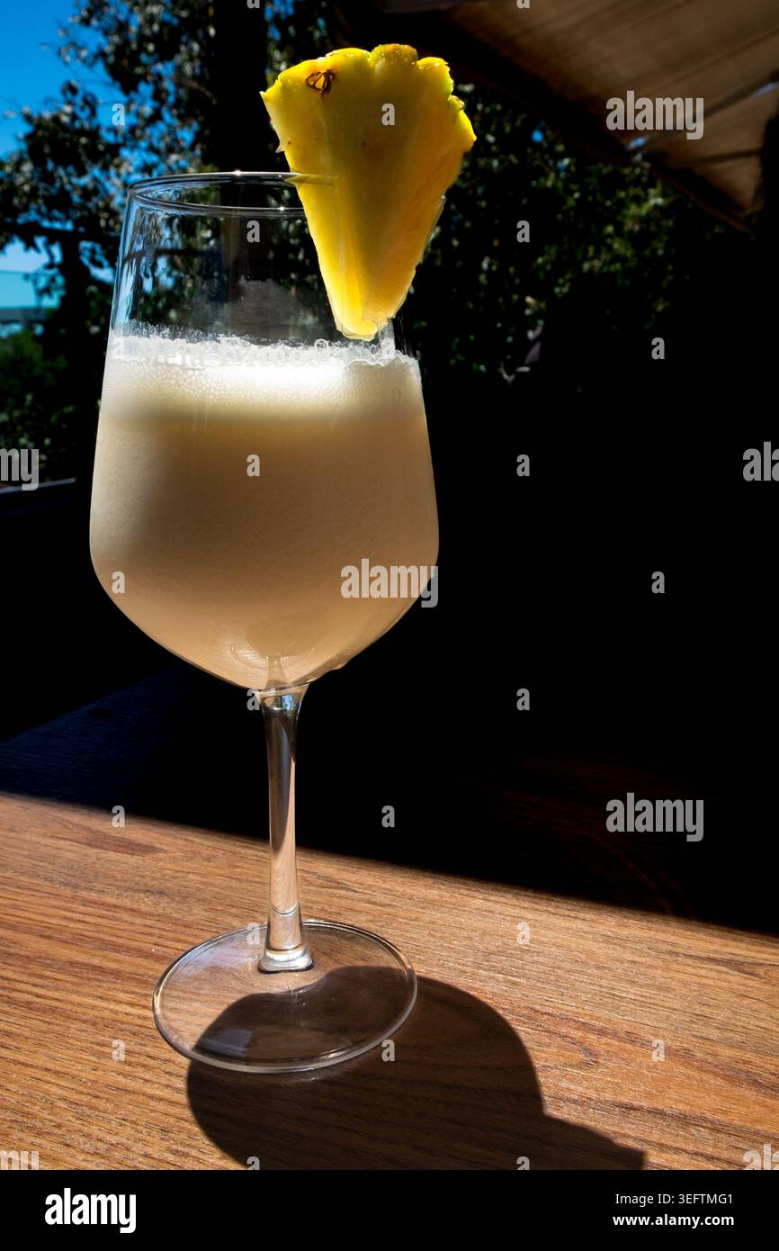Un bicchiere di Pina Colada con una fetta di ananas gialla getta un'ombra su un tavolo di legno. Alberi, cielo blu e tendalino marrone sono visibili nel backgrou Foto Stock