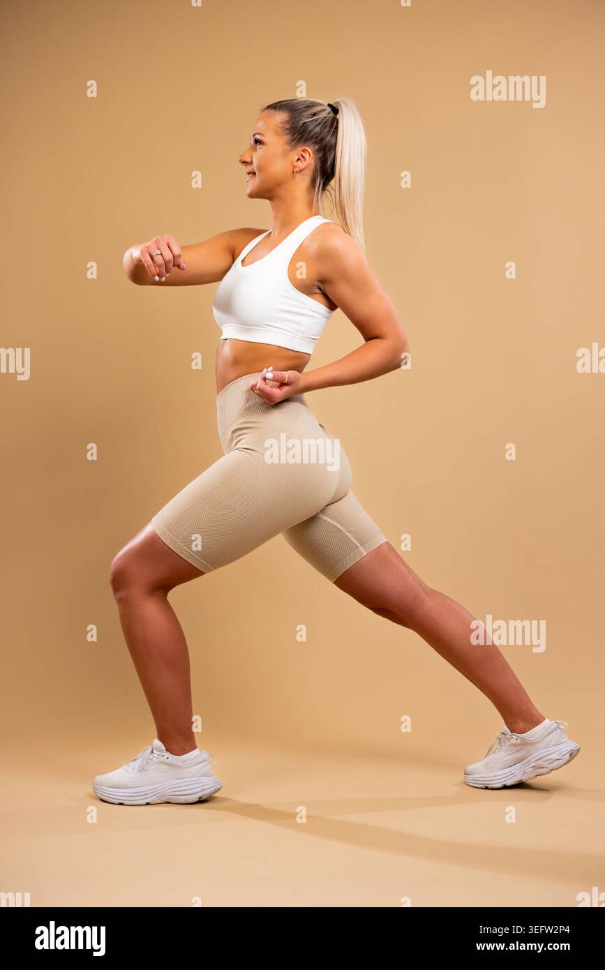 Donna atletica in reggiseno sportivo bianco e leggings beige che esegue affondi in stile studio Foto Stock