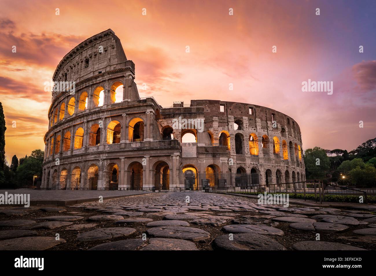 Sorprendente prospettiva a basso angolo che si concentra sulle grandi asfaltatrici in pietra basaltica, conducendo l'occhio verso il Colosseo illuminato sotto un cielo dorato al tramonto. Foto Stock