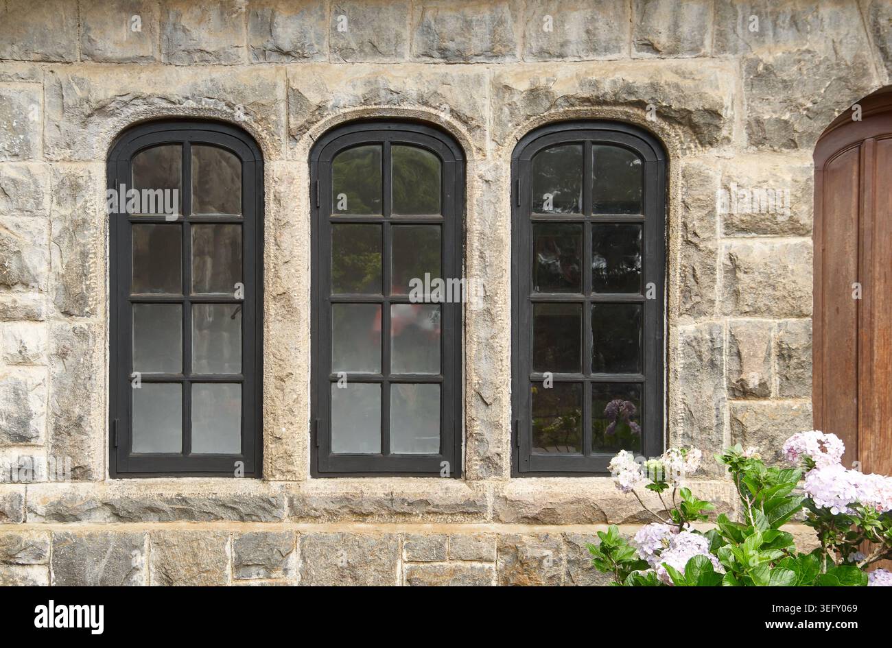 finestre in vetro ad arco nero su pareti rustiche in pietra grigia, telai multi-pannello con vetro riflettente e fiori fioriti, vista laterale dell'europa vintage Foto Stock