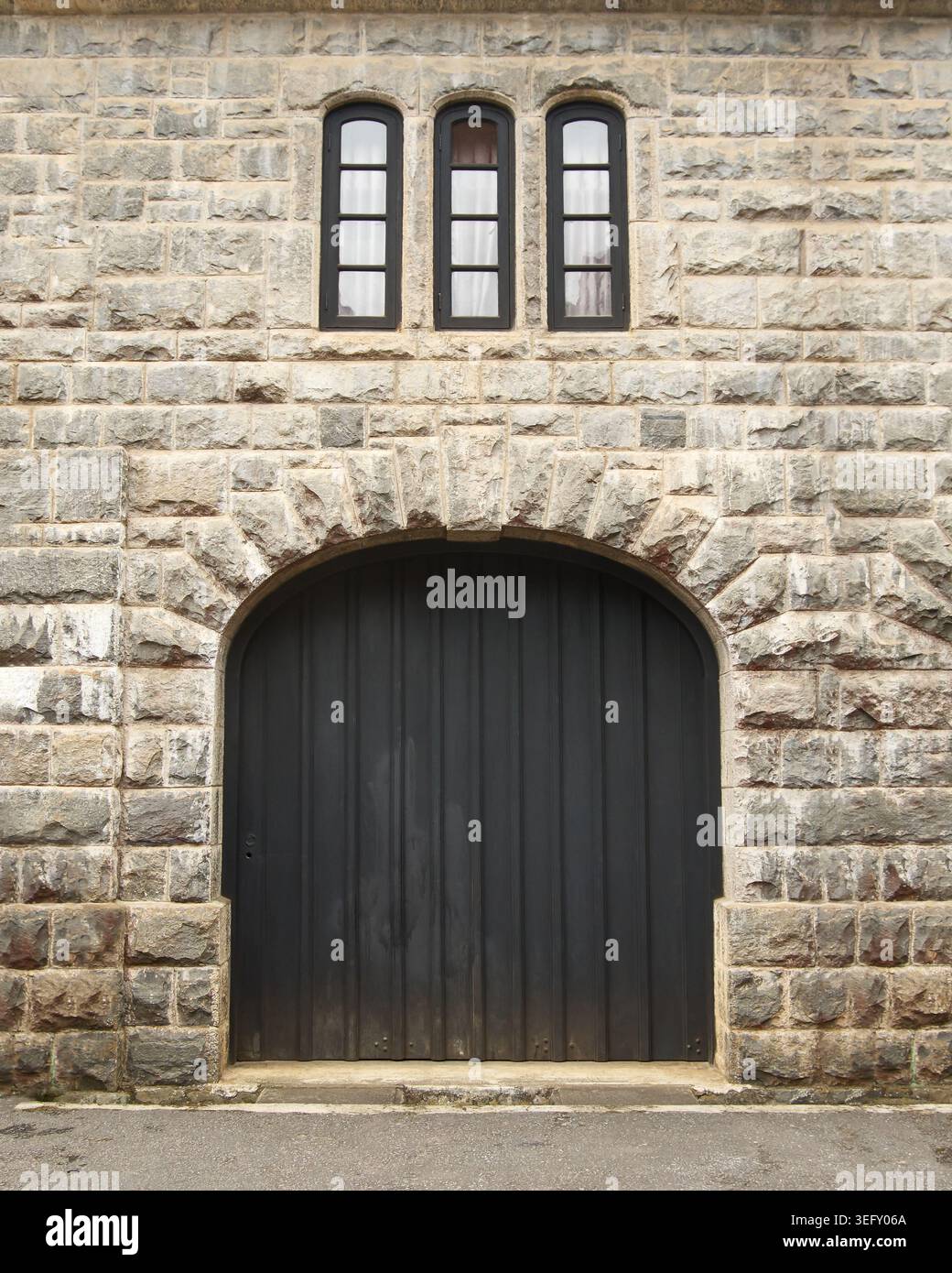 porta ad arco in legno nero su rustico muro in pietra grigia, ingresso simmetrico all'edificio storico con strette finestre, vista laterale del castello medievale Foto Stock