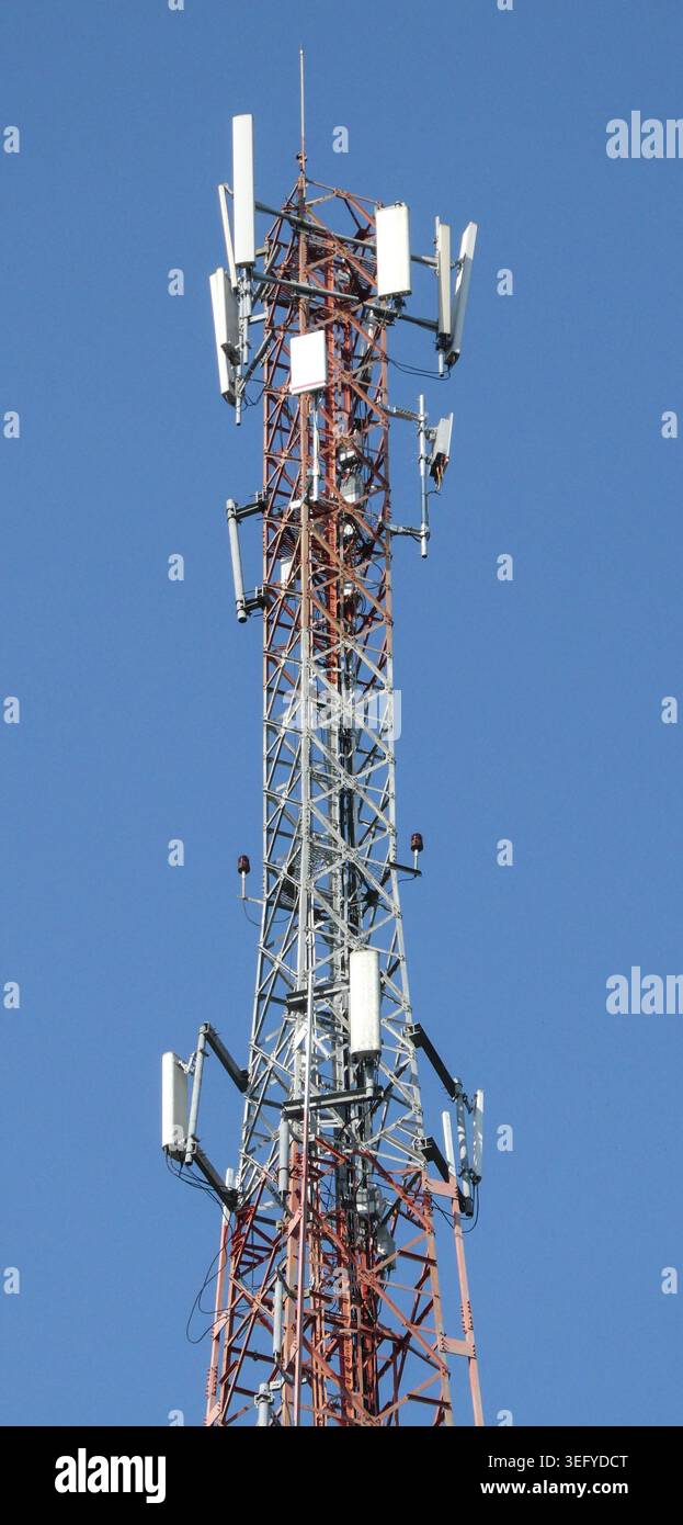 Il montante per telecomunicazioni a reticolo rosso e bianco con antenne a più settori si erge in alto sotto un cielo blu chiaro durante il giorno, trasportando cavi moderni Foto Stock