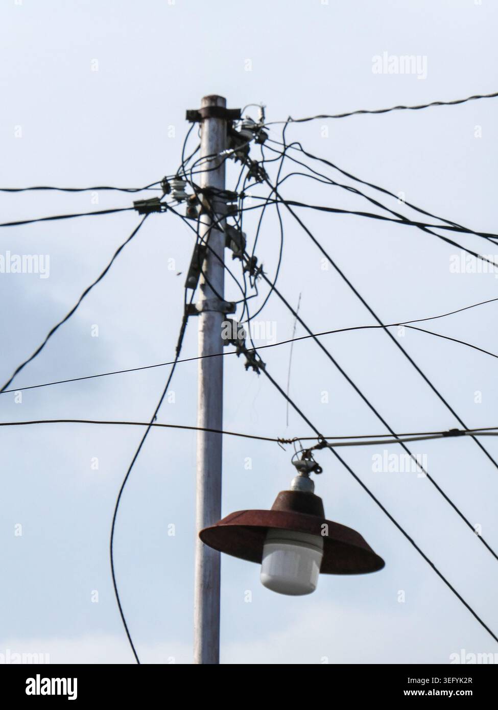 Un'asta resistente agli agenti atmosferici sostiene un groviglio di cavi elettrici e un lampione stradale vintage, sagomato contro un cielo blu brillante durante il giorno in una città Foto Stock