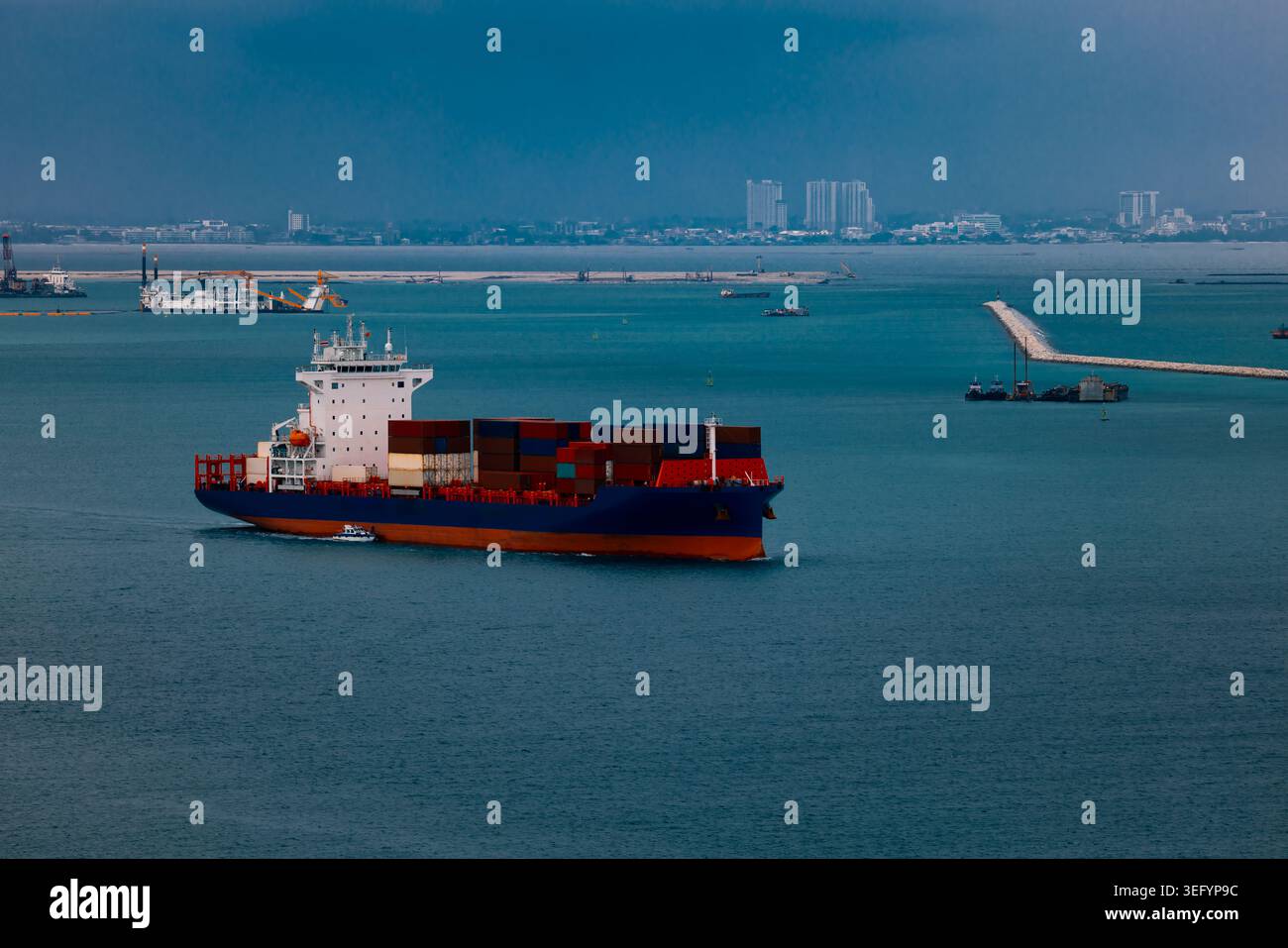 Un cargo commerciale che trasporta container naviga attraverso le acque costiere verso le infrastrutture portuali sotto un cielo soffocato. Foto Stock