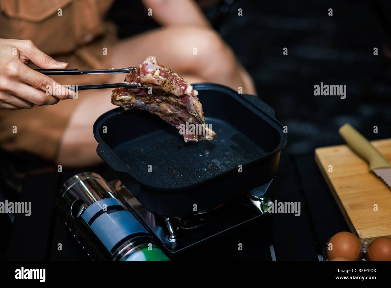 Vista ravvicinata di una persona che usa pinze metalliche per mettere la carne cruda condita in una padella portatile per cucinare all'aperto. Foto Stock