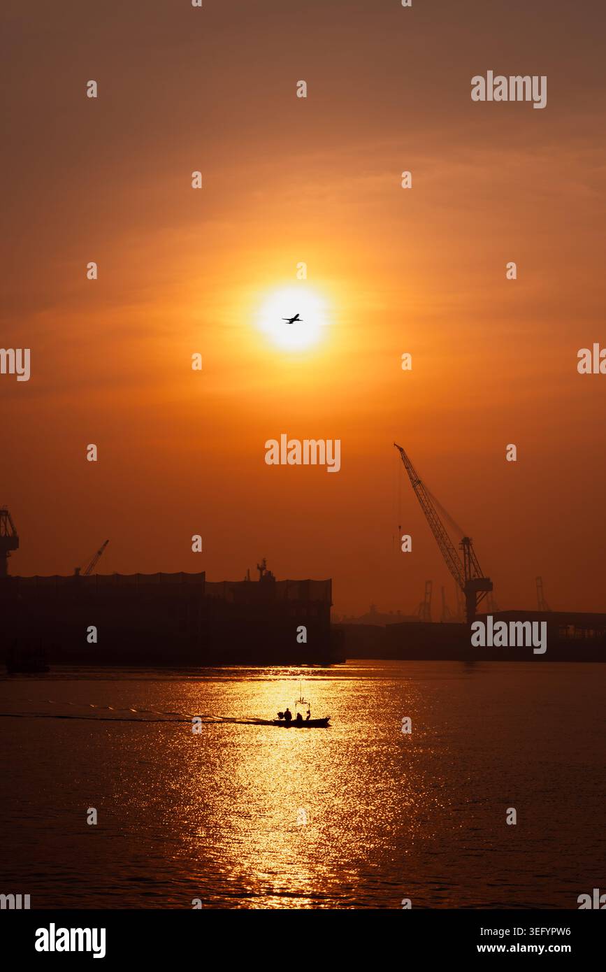 Una sagoma di una piccola nave che attraversa le calme acque del porto durante un vivido tramonto con gru industriali e un aereo commerciale nel cielo dorato. Foto Stock