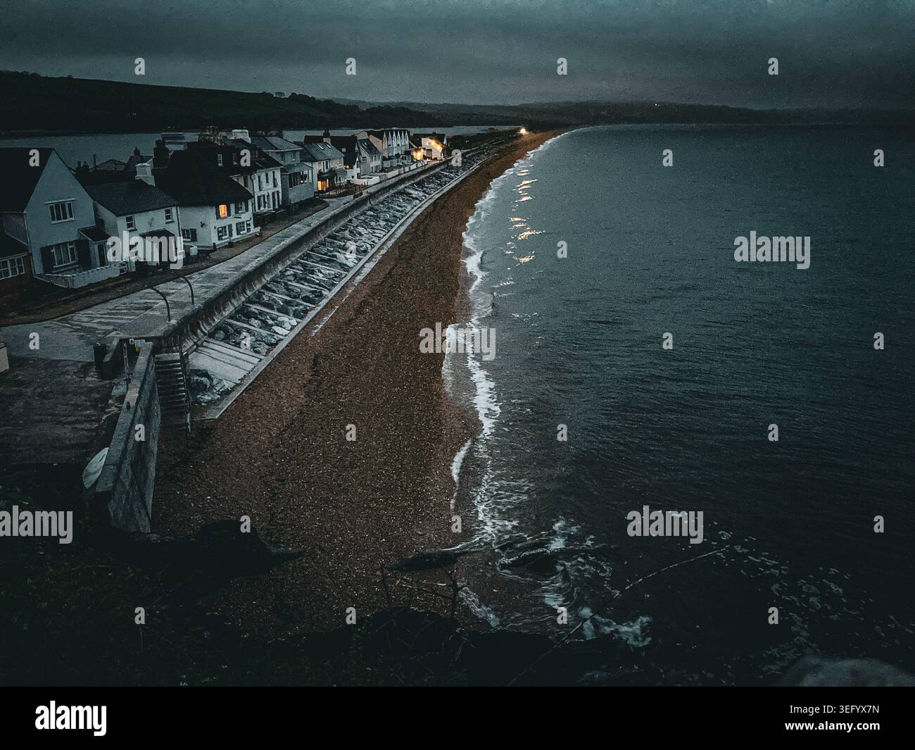 Tramonto a Torcross, Slapton Sands, Devon. Una costa ampia con piccole onde e case sulla lunga spiaggia di ciottoli. - Immagine stock catturata con smartphone