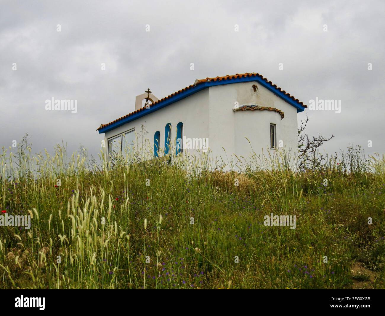 Piccola chiesa bianca tradizionale con una campana su una collina erbosa sotto il cielo nuvoloso. Foto Stock