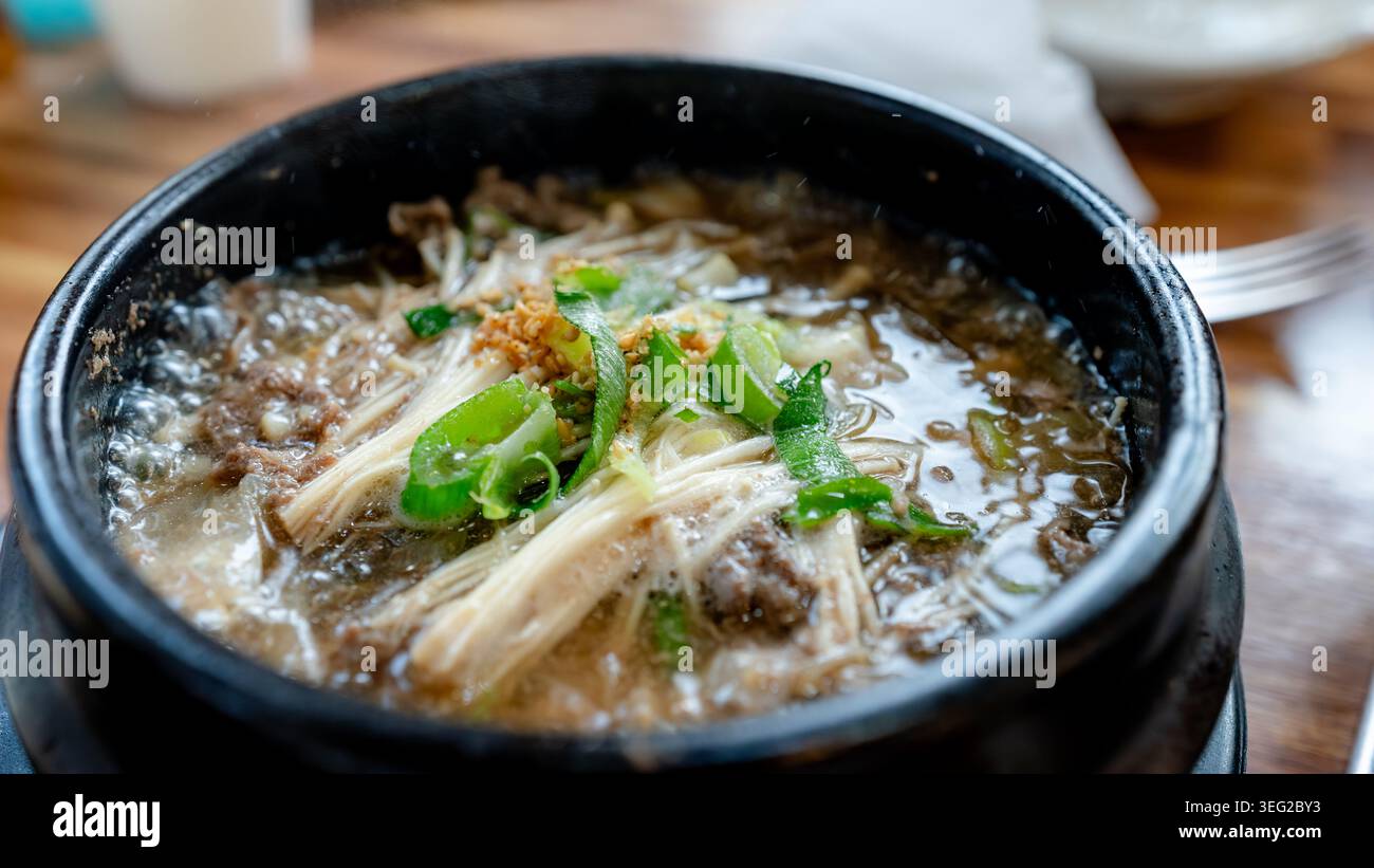 Primo piano di una zuppa coreana bulgogi fumante servita in una pentola di pietra nera, brodo bollente con carne di manzo, funghi, scalleone e condimento con sesamo su una Foto Stock