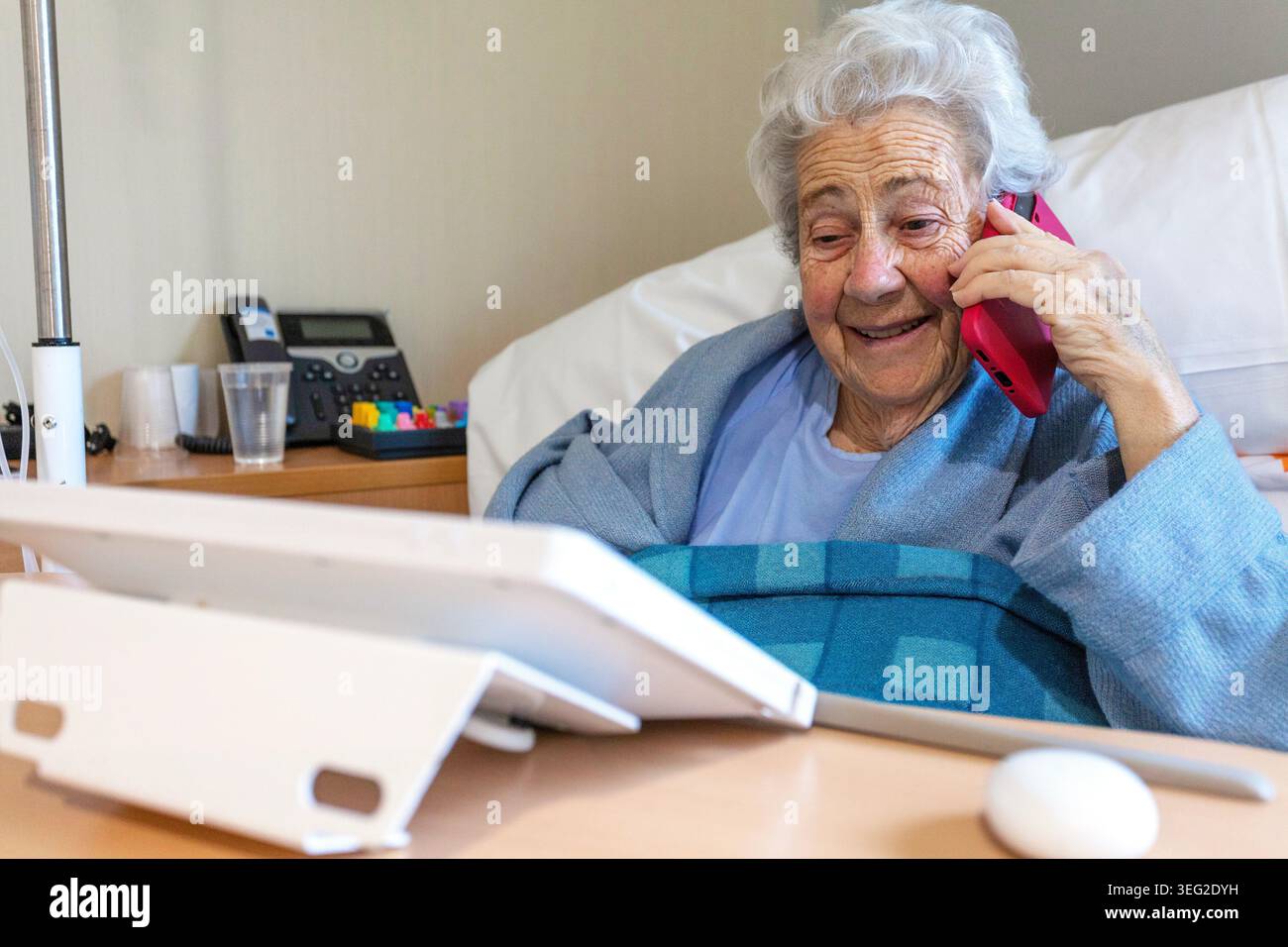 Una donna anziana in un letto d'ospedale sorride calorosamente mentre parla al telefono. Con un tablet e attrezzature mediche nelle vicinanze, appare contenuto e comfo Foto Stock