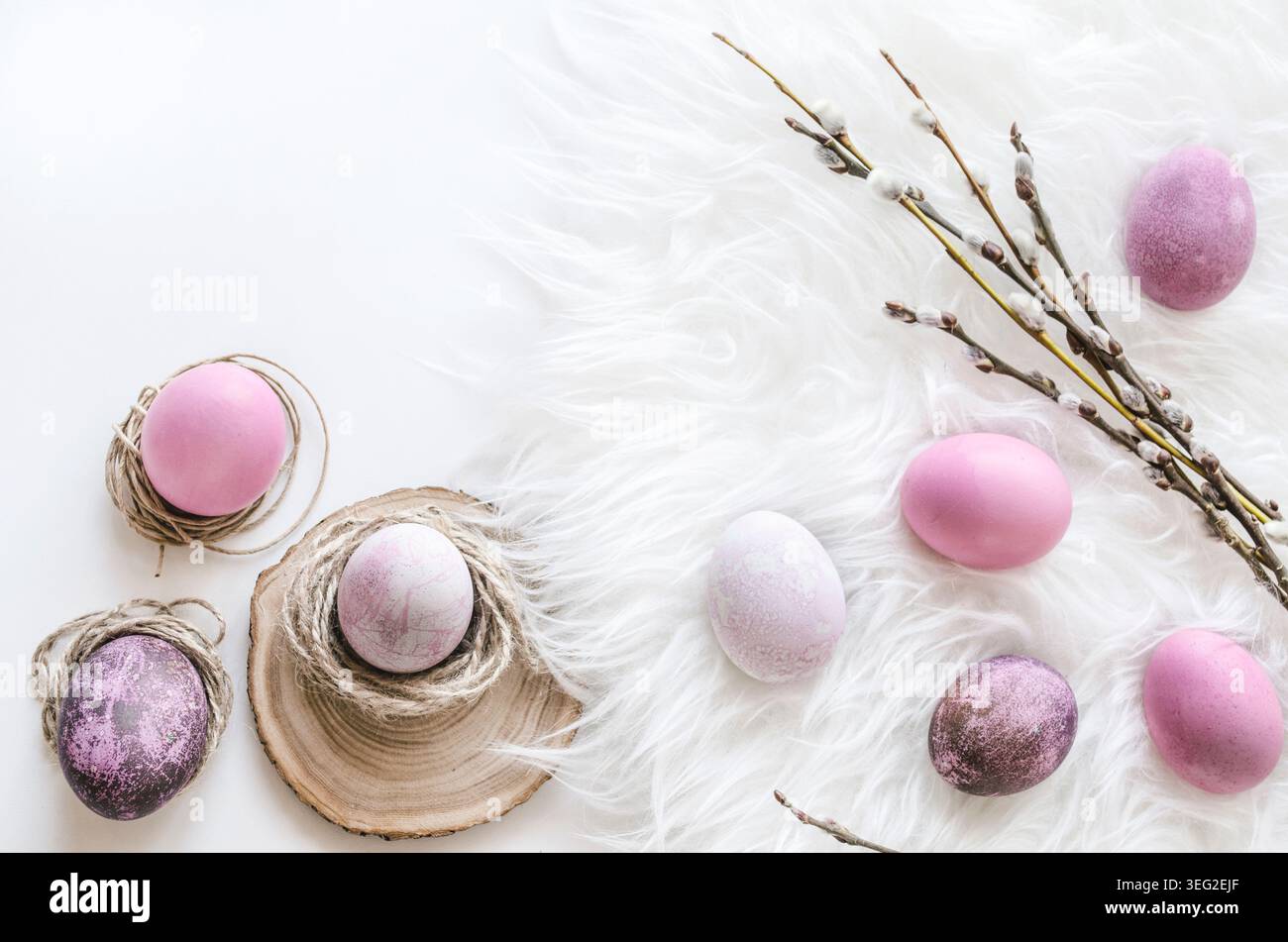 Composizione pasquale minimale con uova viola pastello e rosa disposte su morbida pelliccia bianca, annidate in nidi naturali e abbinate a una crusca primaverile in erba Foto Stock