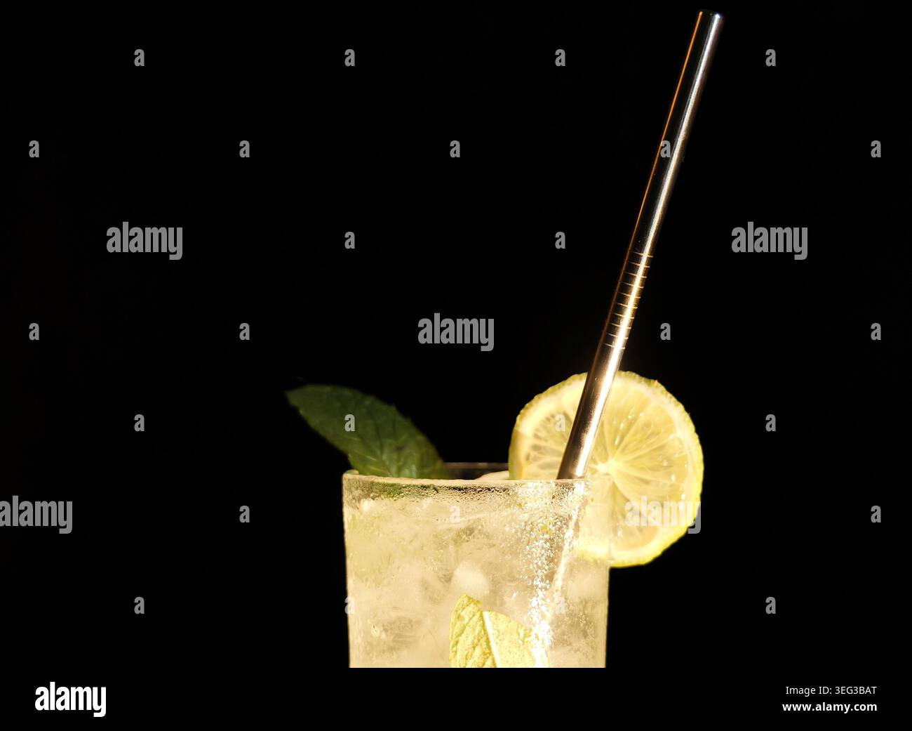 Cocktail ghiacciato con fette di lime fresche, foglie aromatiche di menta e un'elegante cannuccia riutilizzabile in metallo, presentata in un bicchiere su sfondo scuro Foto Stock