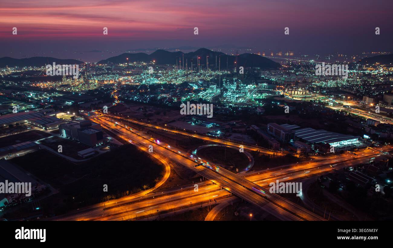 Una vista ad alto angolo di una raffineria di petrolio illuminata e di un trafficato interscambio autostradale con sentieri luminosi sotto un vivace cielo crepuscolo. Foto Stock