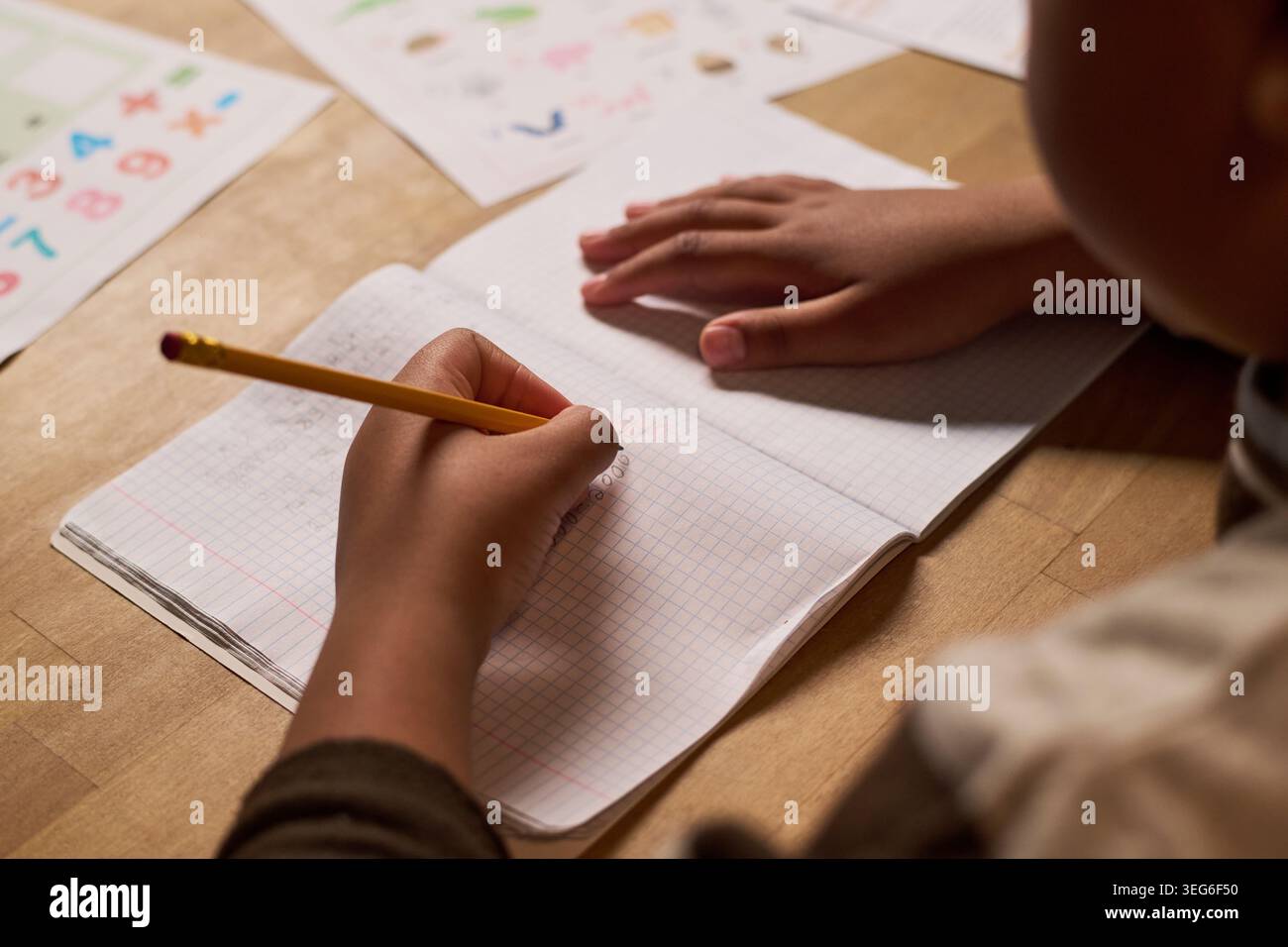 Primo piano di un bambino che scrive su un notebook con una matita, pratica i numeri e le prime abilità matematiche a tavola. Concetto educativo per la homeschooling, il tutoring, i materiali per l'apprendimento in classe Foto Stock