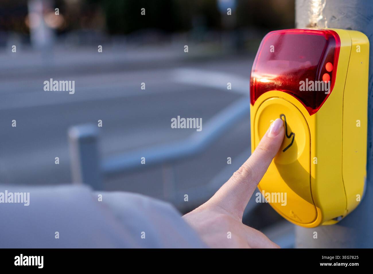 Una persona preme il pulsante del segnale stradale giallo in corrispondenza di un passaggio pedonale. Il sole sta tramontando e i veicoli passano sulla strada accanto al marciapiede. Foto Stock