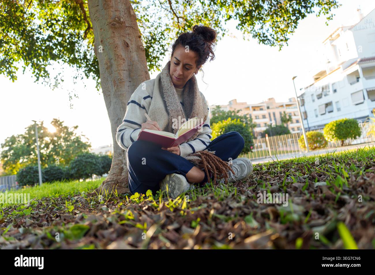 Donna che scrive su un diario mentre siede sull'erba sotto un albero, godendo momenti riflessivi in uno spazio verde urbano Foto Stock