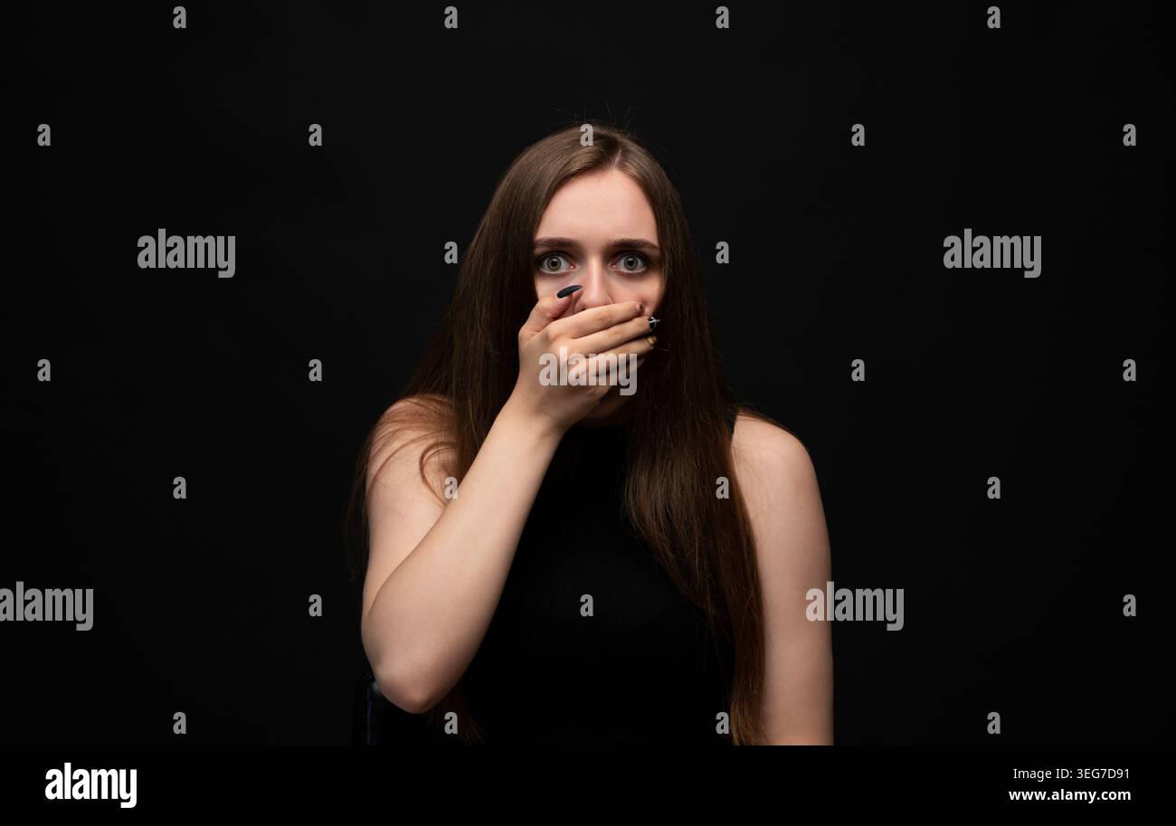 Giovane donna che si copre la bocca con la mano, esprimendo shock e paura con gli occhi spalancati su uno sfondo scuro Foto Stock