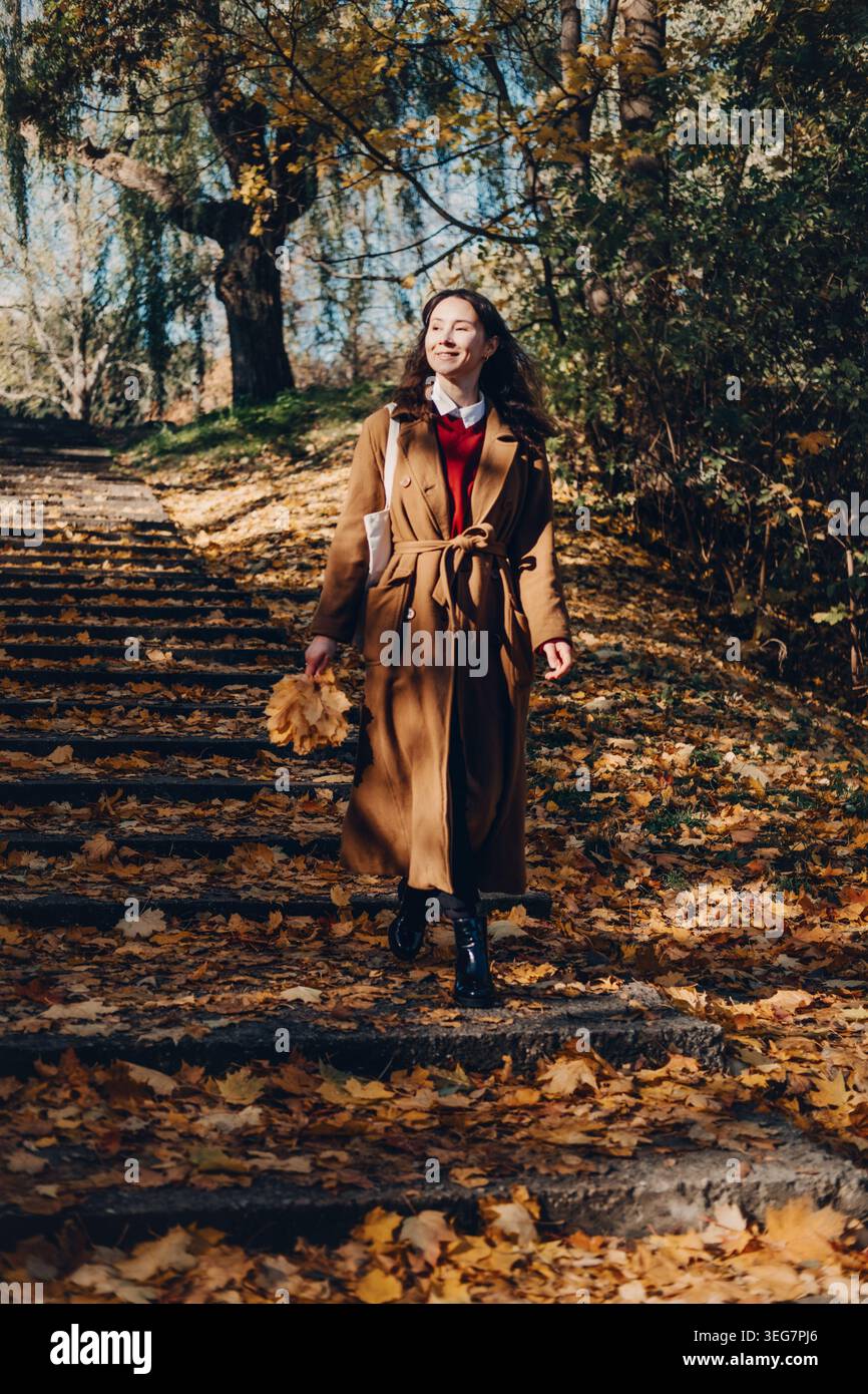 Una donna cammina lungo i gradini di pietra nel parco autunnale tenendo le foglie. Celebra i cambiamenti stagionali autunnali, l'ottimismo e il benessere, autentiche espressioni felici. Foto Stock