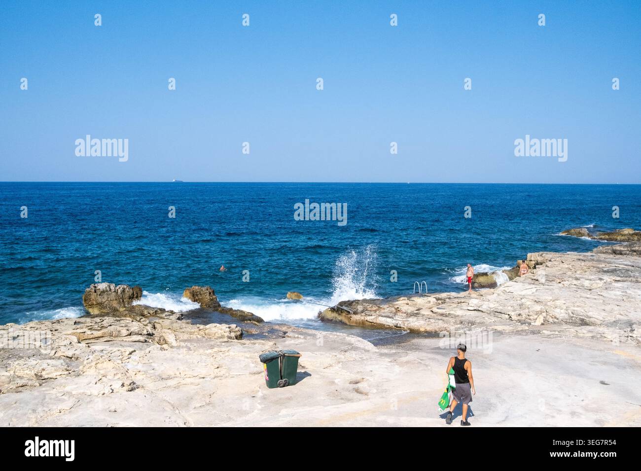 Vista della costa rocciosa della baia di Rinella con il Mar Mediterraneo, le onde e un pedone che trasporta oggetti da spiaggia a Kalkara, regione sud-orientale, M Foto Stock
