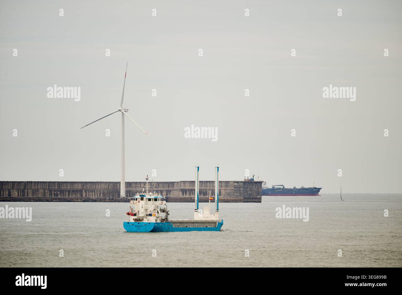 Nave da carico che naviga nelle acque portuali con una turbina eolica che fornisce energia pulita e altre navi all'orizzonte remoto Foto Stock