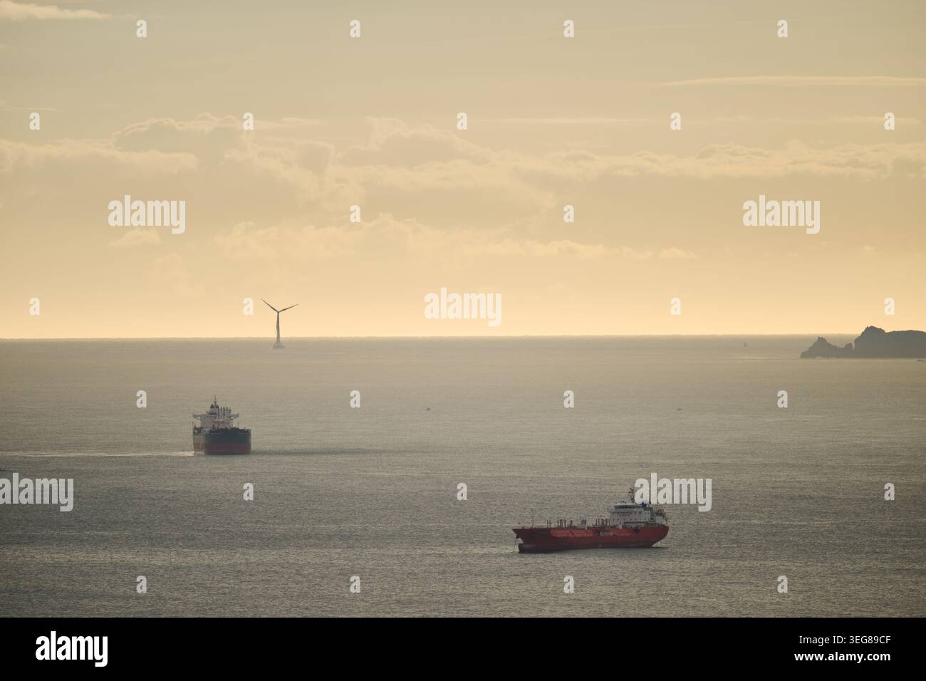 Due navi commerciali navigano in un mare calmo con una turbina eolica offshore che genera energia rinnovabile contro un cielo nebuloso Foto Stock