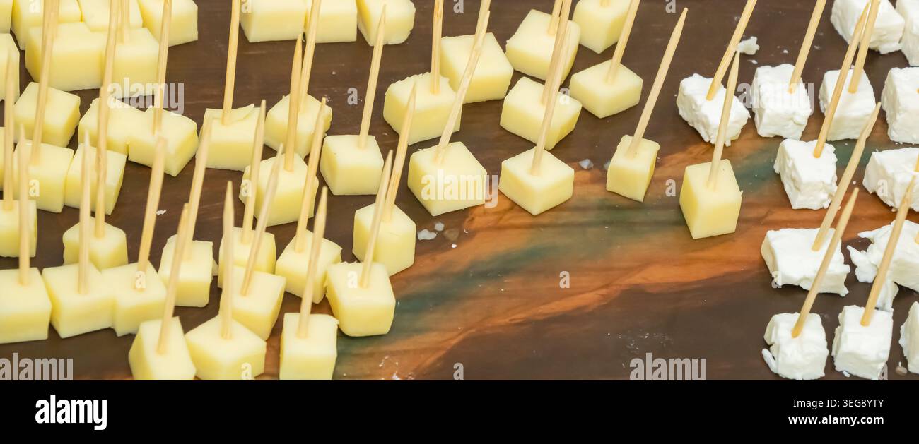 Cubetti di formaggio giallo e bianco assortiti con stuzzicadenti in legno esposti su un banco di servizio rustico durante la degustazione, il buffet o l'evento di catering. Foto Stock