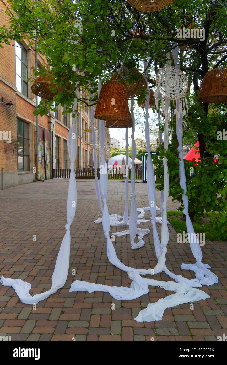 Elegante decorazione per eventi in stile boho con nastri in tessuto bianco appesi, sognatori e lampade in vimini sotto rami di alberi verdi all'aperto Foto Stock