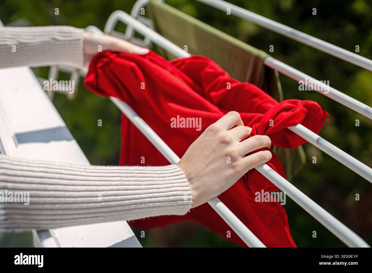 Appendere abiti rossi sul rack di asciugatura del balcone. Mani che appendono un indumento rosso brillante su una griglia di asciugatura da balcone all'aperto in condizioni di sole. Foto Stock