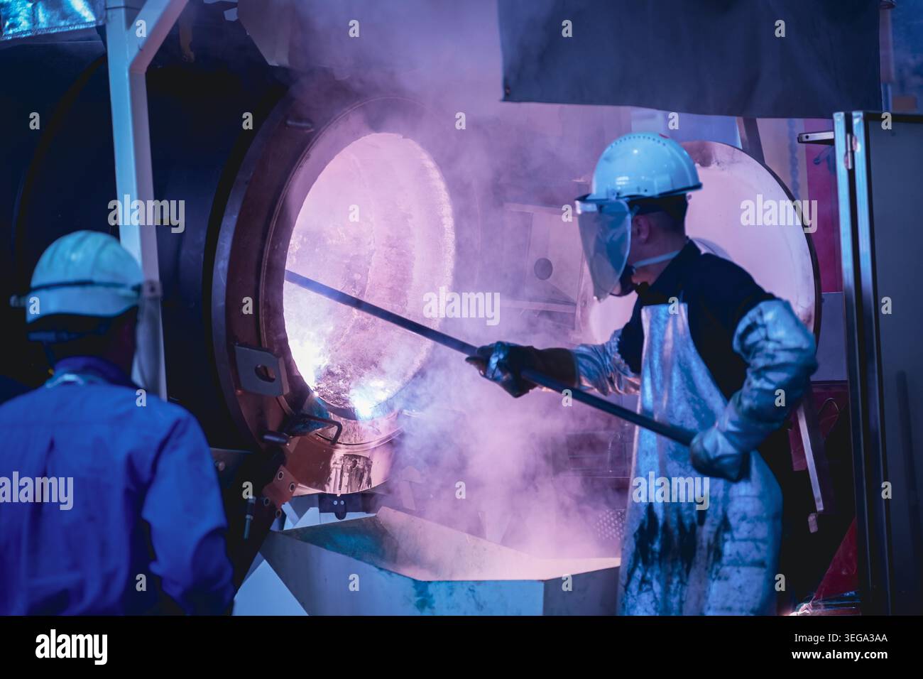 Un lavoratore con un equipaggiamento di protezione completo, tra cui uno schermo facciale e una tuta resistente al calore, utilizza un'asta per sondare all'interno di un forno da fonderia caldo. Foto Stock