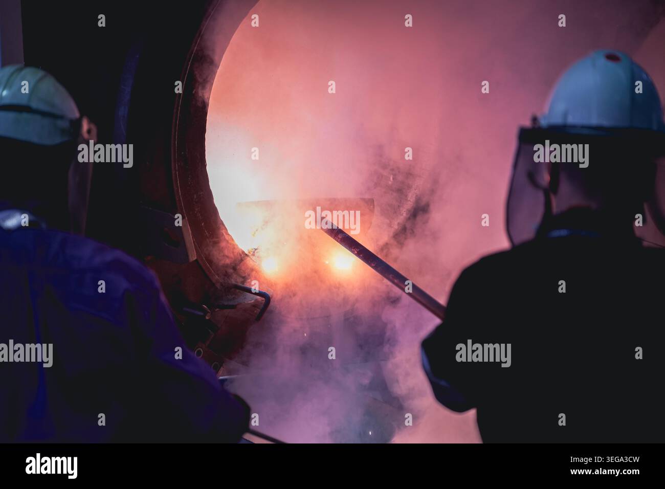 Un lavoratore con un equipaggiamento di protezione completo, tra cui uno schermo facciale e una tuta resistente al calore, utilizza un'asta per sondare all'interno di un forno da fonderia caldo. Foto Stock