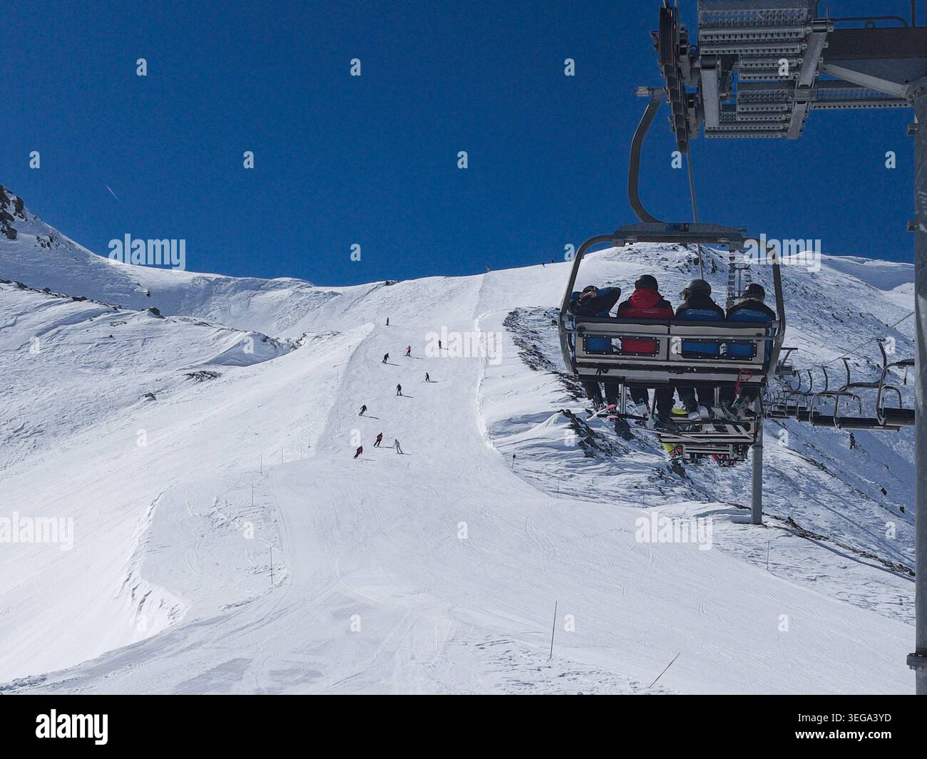 Una scena invernale a la Thuile, che mostra una seggiovia per quattro persone che sale su una pista innevata, con gli sciatori che scendono lungo la pista ben curata sotto un cielo limpido Foto Stock