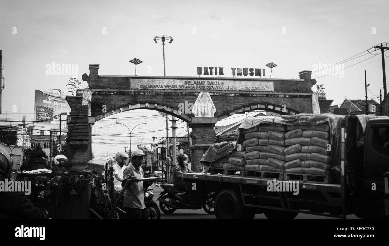 Fotografia di strada monocromatica che cattura l'iconica arcata in mattoni dell'area turistica di Batik Trusmi a Cirebon, Giava occidentale, Indonesia. La scena raffigura il traffico mattutino con un camion a pianale che trasporta sacchi di merci, motociclisti locali e pedoni che passano, creando un'atmosfera grintosa e autentica del commercio e della vita quotidiana locale. Foto Stock