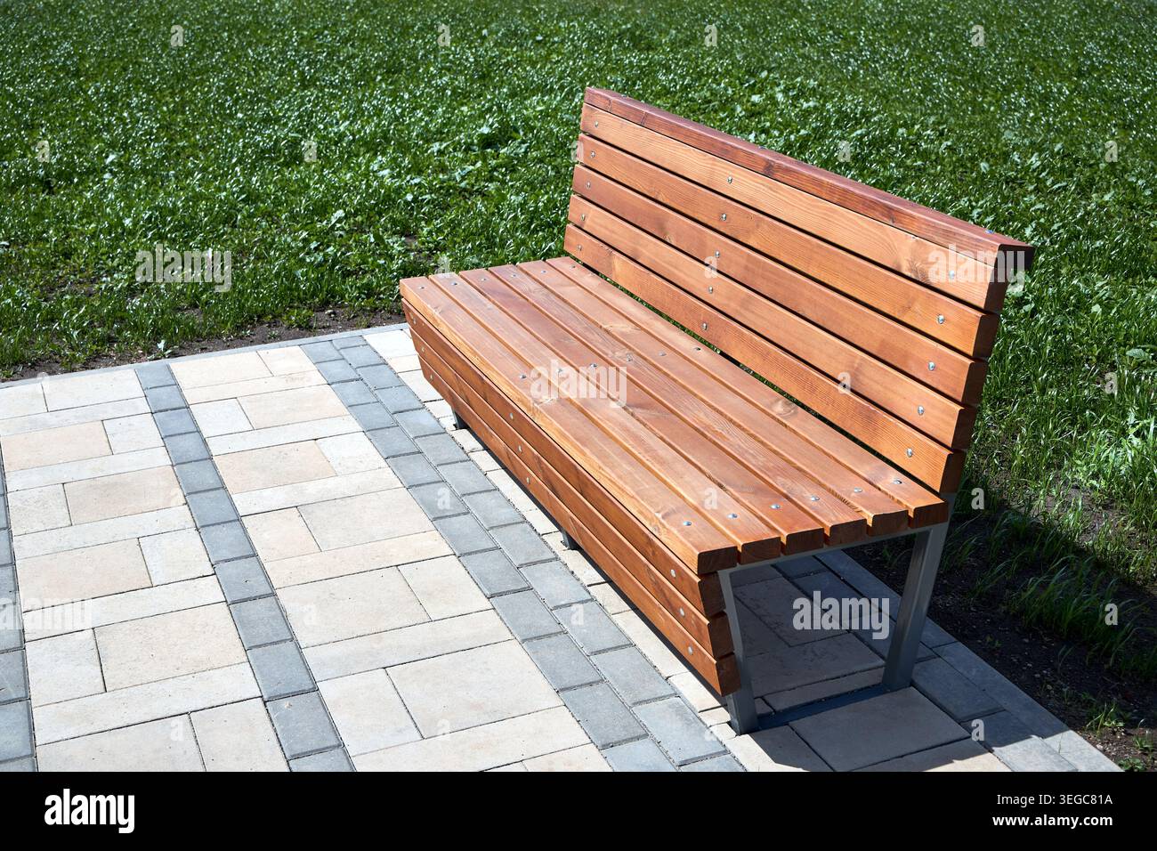 La panchina in legno si erge su un sentiero lastricato in pietra accanto al prato verde. I moderni mobili da esterno presentano tavole in legno e gambe in metallo. La giornata di sole crea un'atmosfera elegante Foto Stock