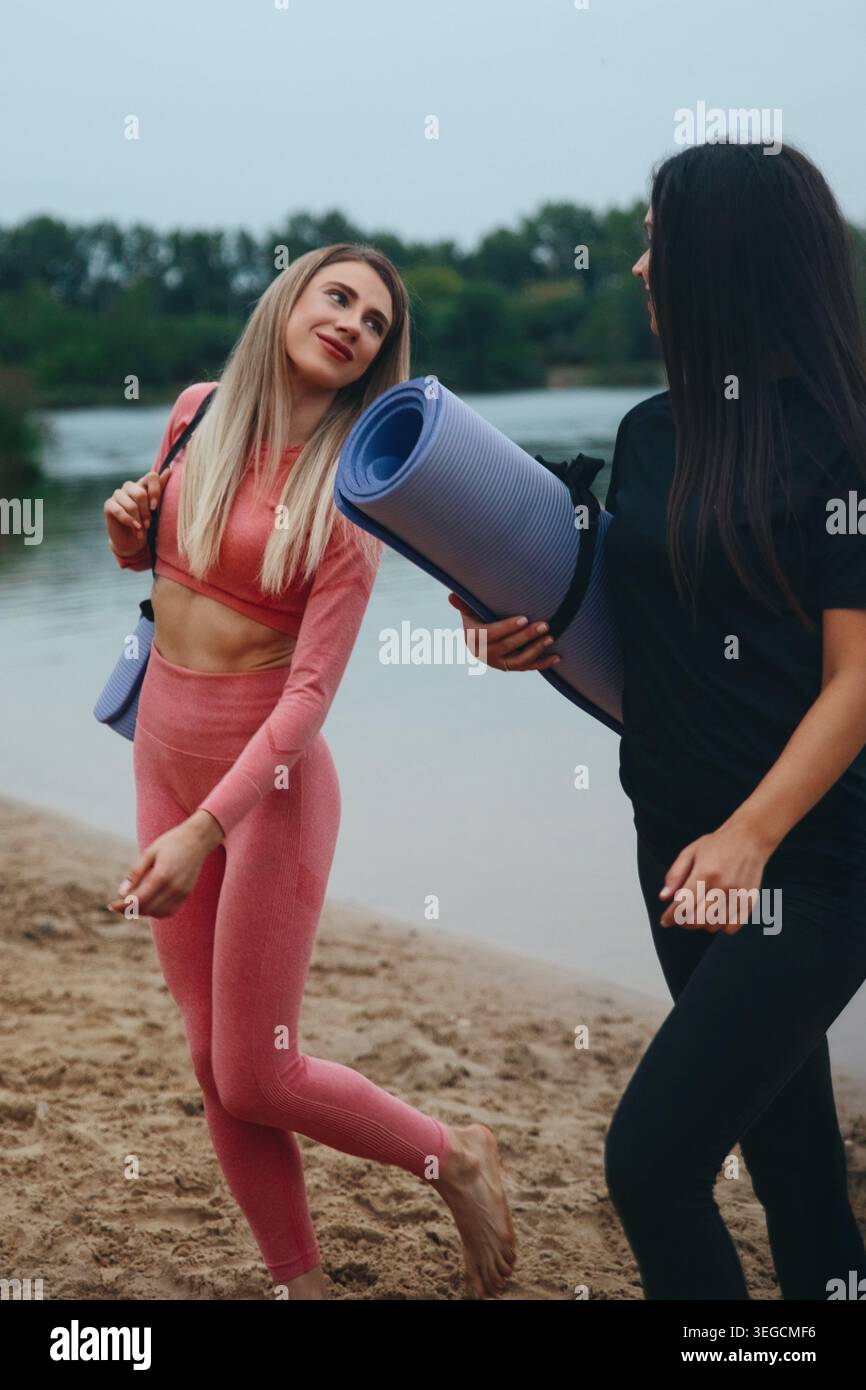 Due donne camminano lungo la riva sabbiosa del lago portando con sé stuoie per lo yoga, sorridendo e chiacchierando sul loro percorso di fitness in una splendida giornata. Foto Stock