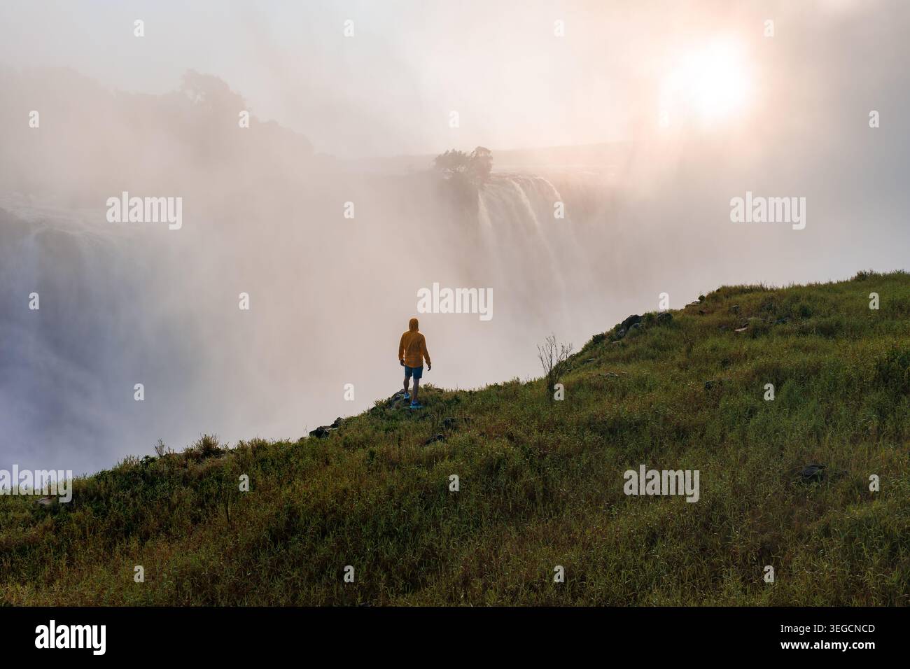 La persona in giacca gialla guarda le cascate delle cascate Victoria Falls al tramonto Foto Stock