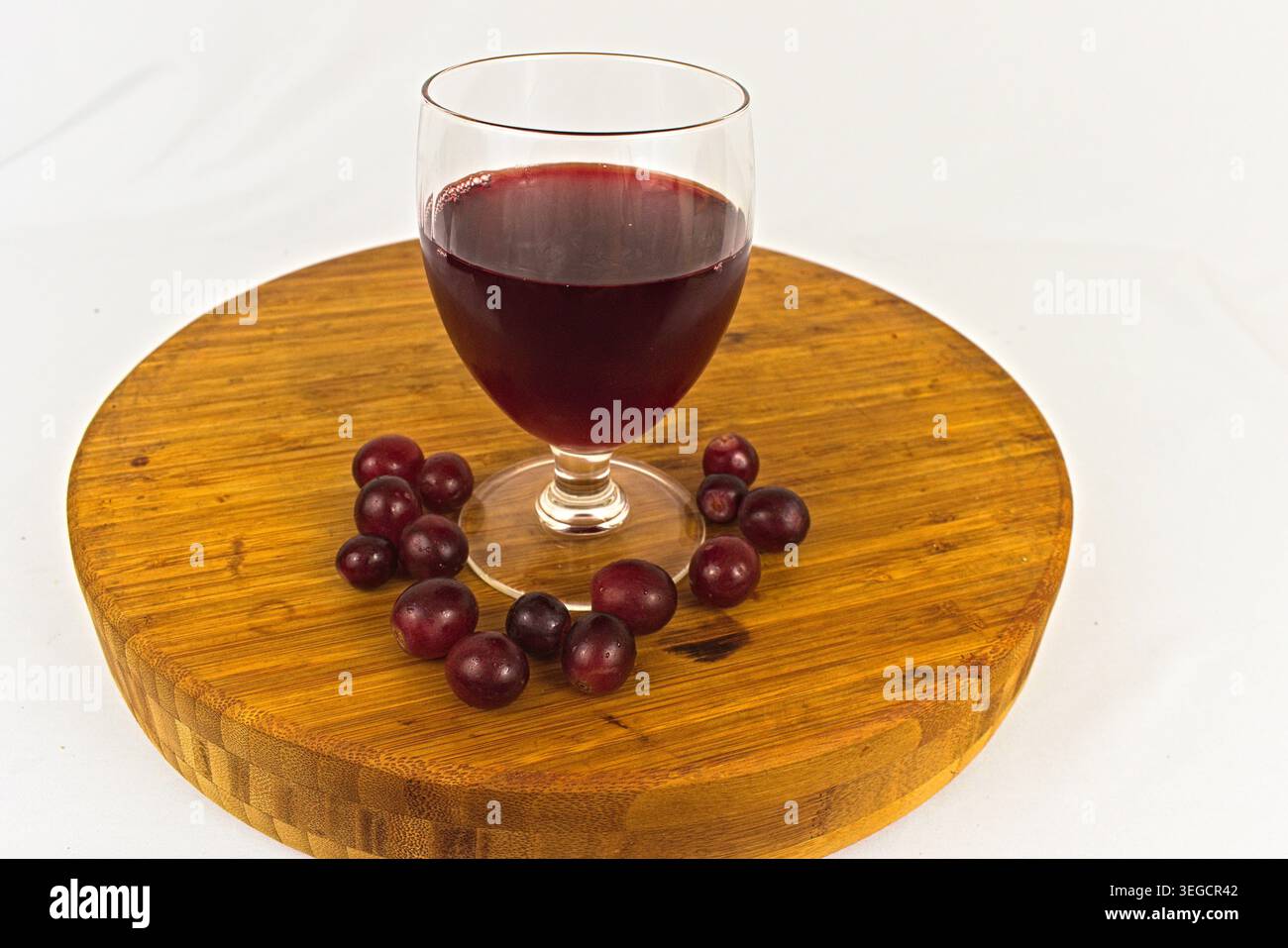 Bicchiere di succo d'uva rosso intenso circondato da uva intera fresca su un tagliere di legno. Foto Stock