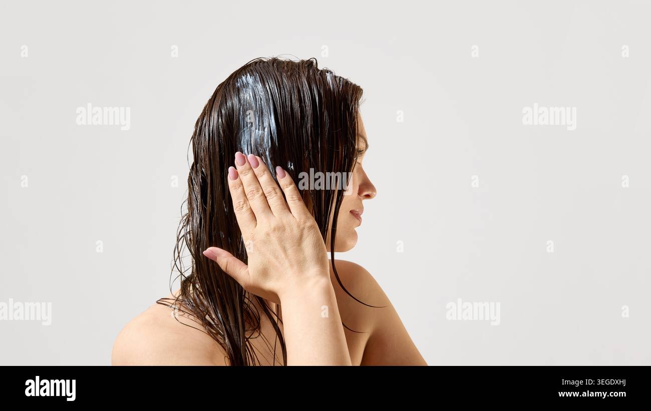 Donna che applica la maschera per capelli ai capelli della bruna bagnati su un pulito sfondo studio. Foto Stock