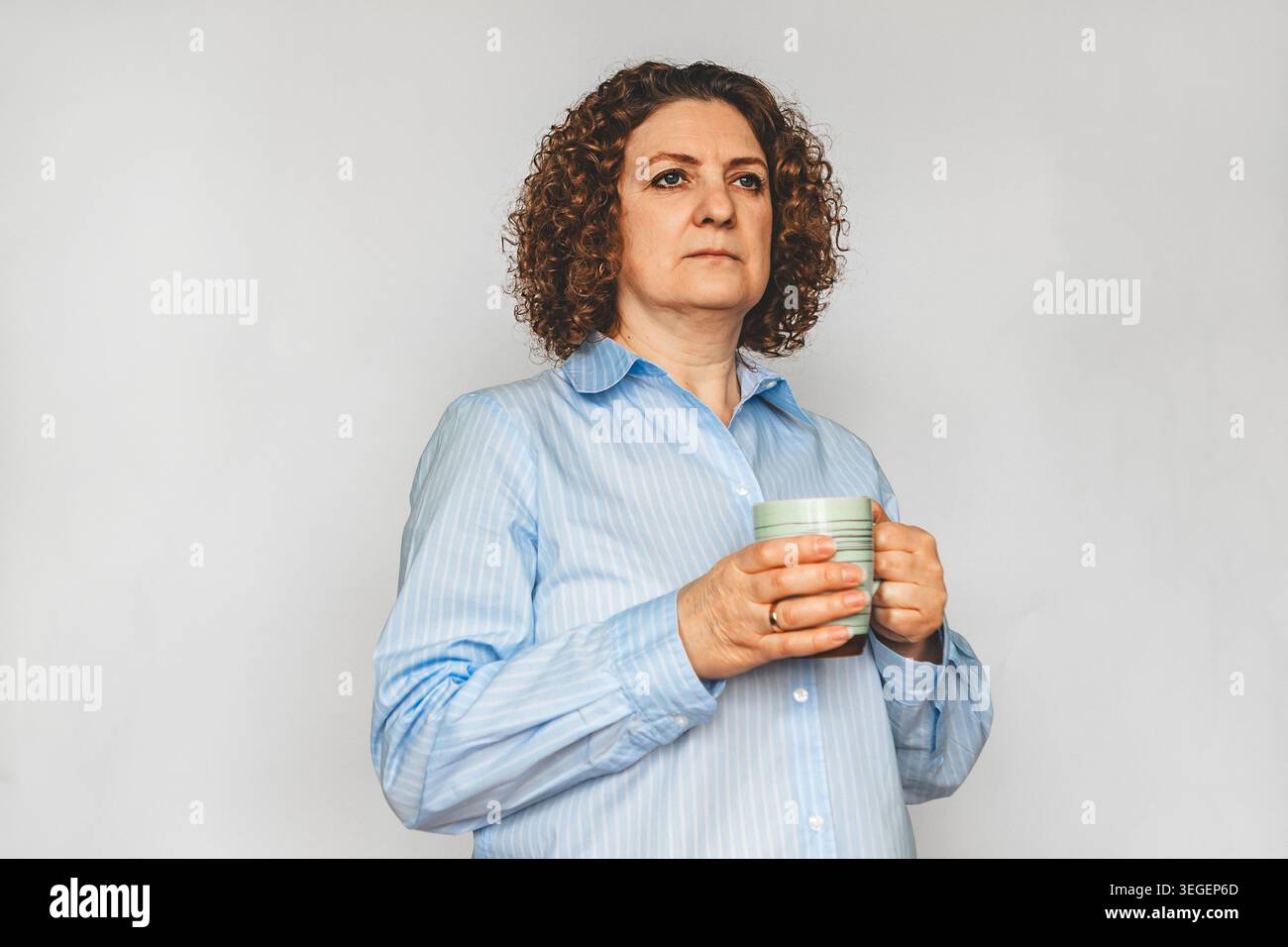 Donna di mezza età premurosa che tiene in mano una tazza e guarda in lontananza con una camicia blu Foto Stock