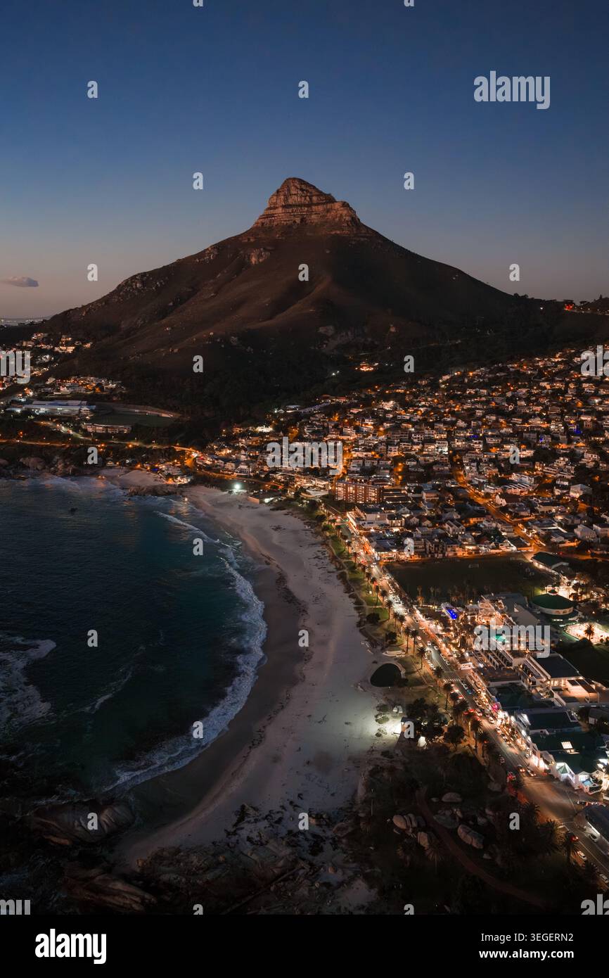La scena aerea mostra Lion's Head sopra Camps Bay e Clifton a città del Capo. I semafori tracciano strade tortuose, spiagge pallide si incontrano per il surf e una S blu viola Foto Stock