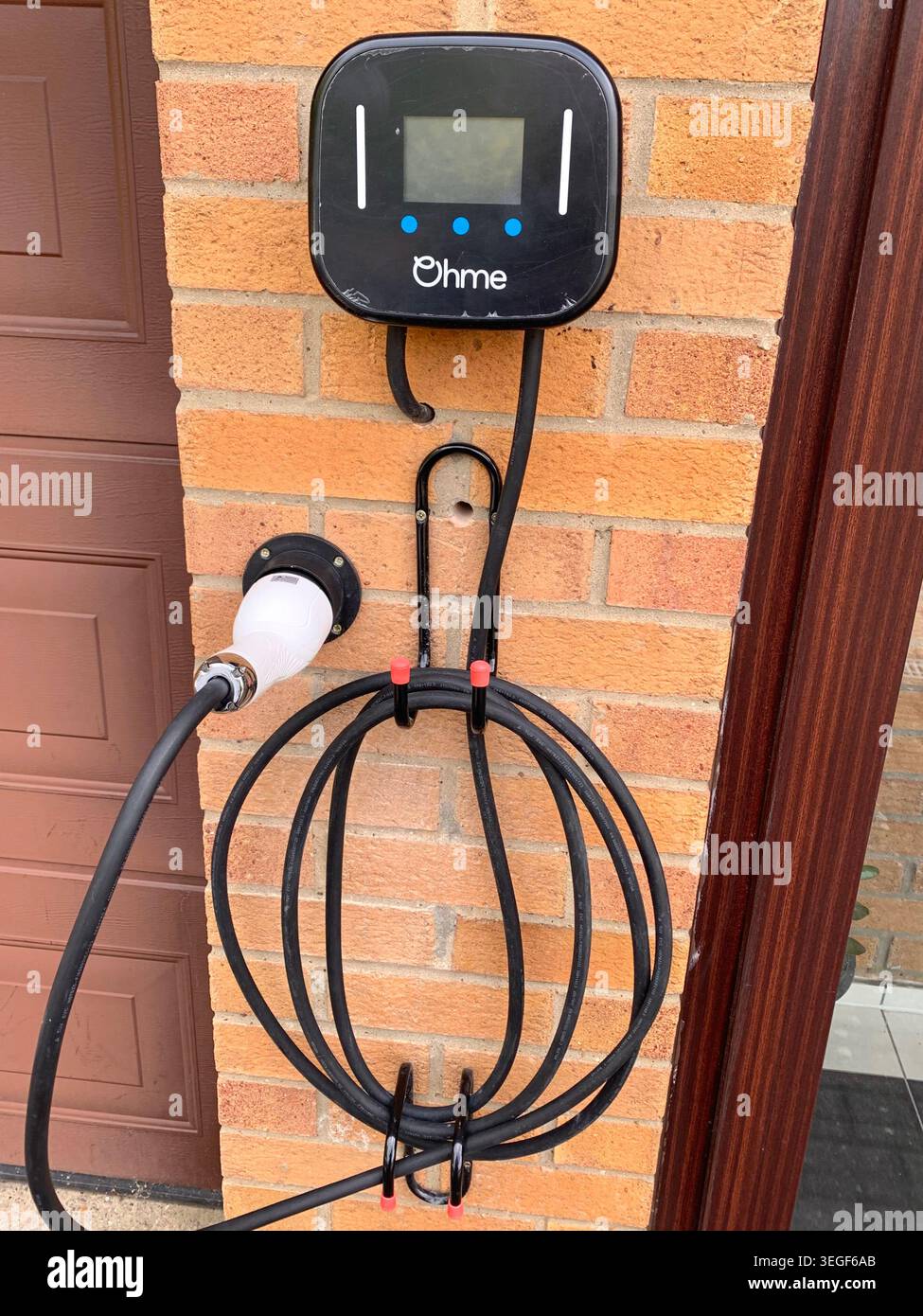 Caricabatterie per auto elettriche Ohme montato su una parete in mattoni con cavi ben avvolti accanto alla porta di un garage a Northampton, Northamptonshire, Inghilterra, Regno Unito - Immagine stock catturata con smartphone