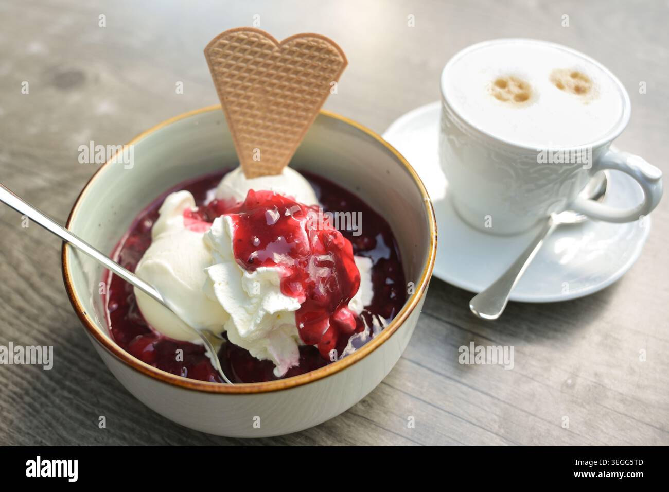 Sundae, palline di gelato alla vaniglia con panna montata e composta di frutta rossa in una ciotola e una tazza di cappuccino su un tavolo di legno, dessert estivo, sel Foto Stock